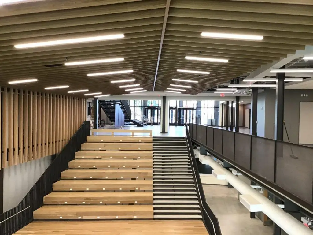 Modern multi-level interior with tiered wooden seating, a staircase, railings, and a slatted ceiling with linear lights