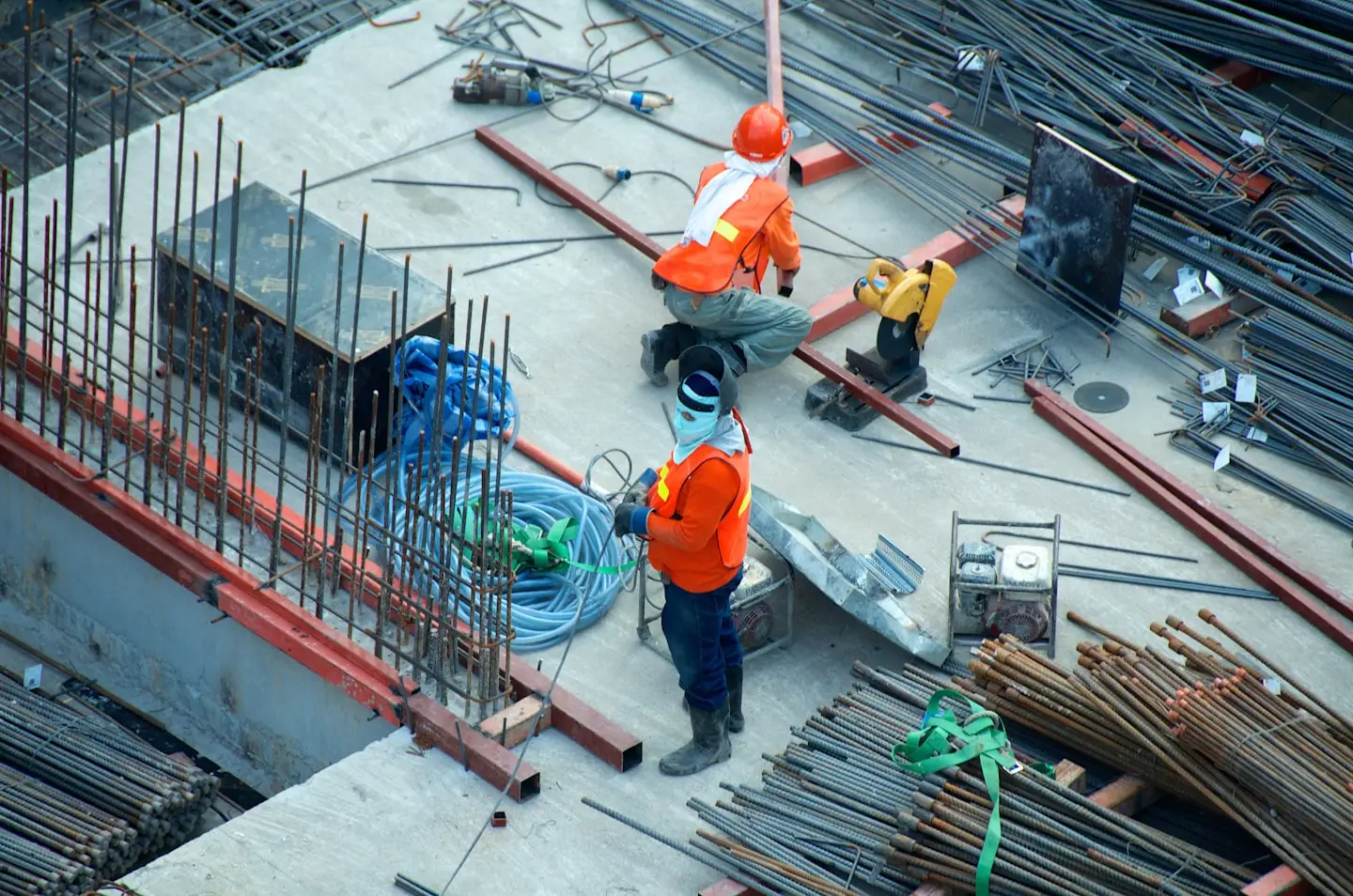 Two construction workers in orange safety clothing work among steel reinforcing bars on a concrete deck.