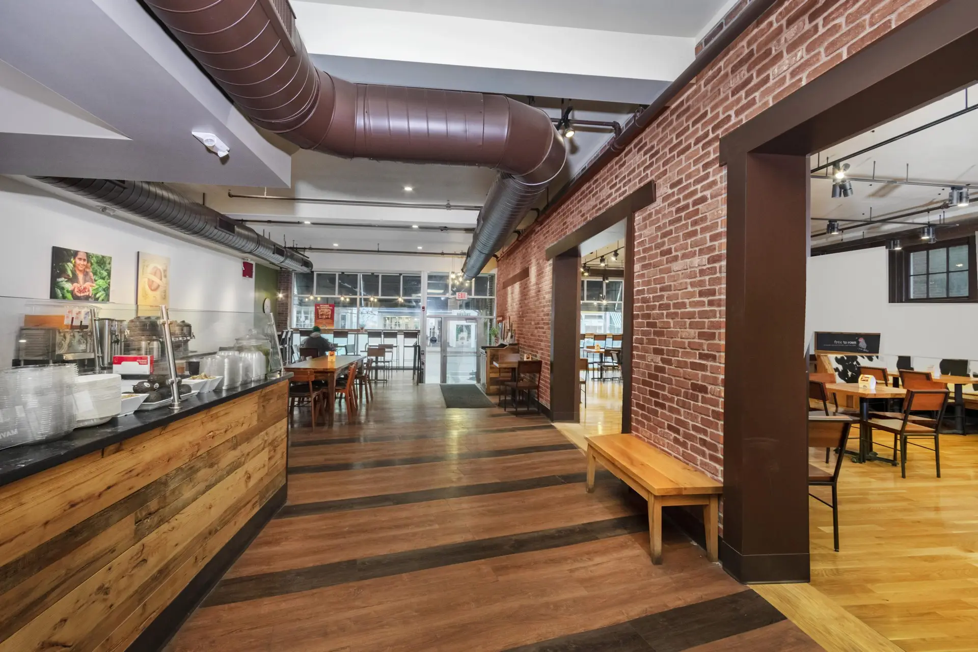 Interior view of a caf&eacute; with a wood-paneled counter, exposed ductwork, brick walls, tables, and chairs.