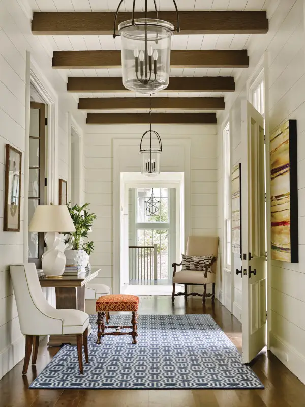 Bright white-paneled hallway with exposed wood ceiling beams, pendant lights, seating, artwork, and a patterned rug.