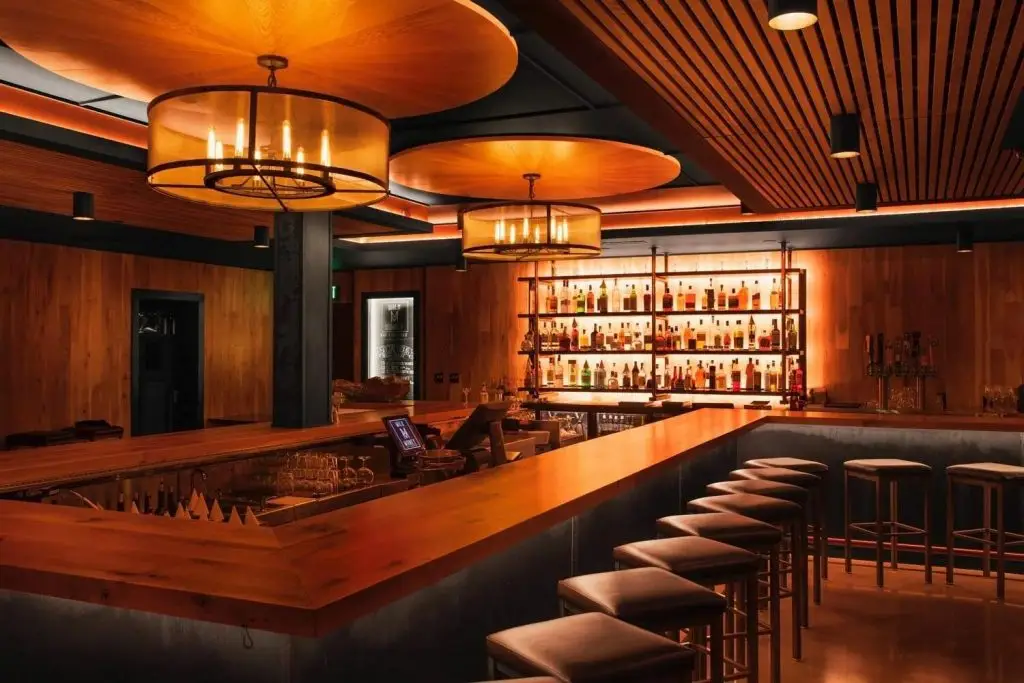 Interior of a warmly lit bar with a long wooden counter, stools, pendant lights, and shelves of liquor bottles.