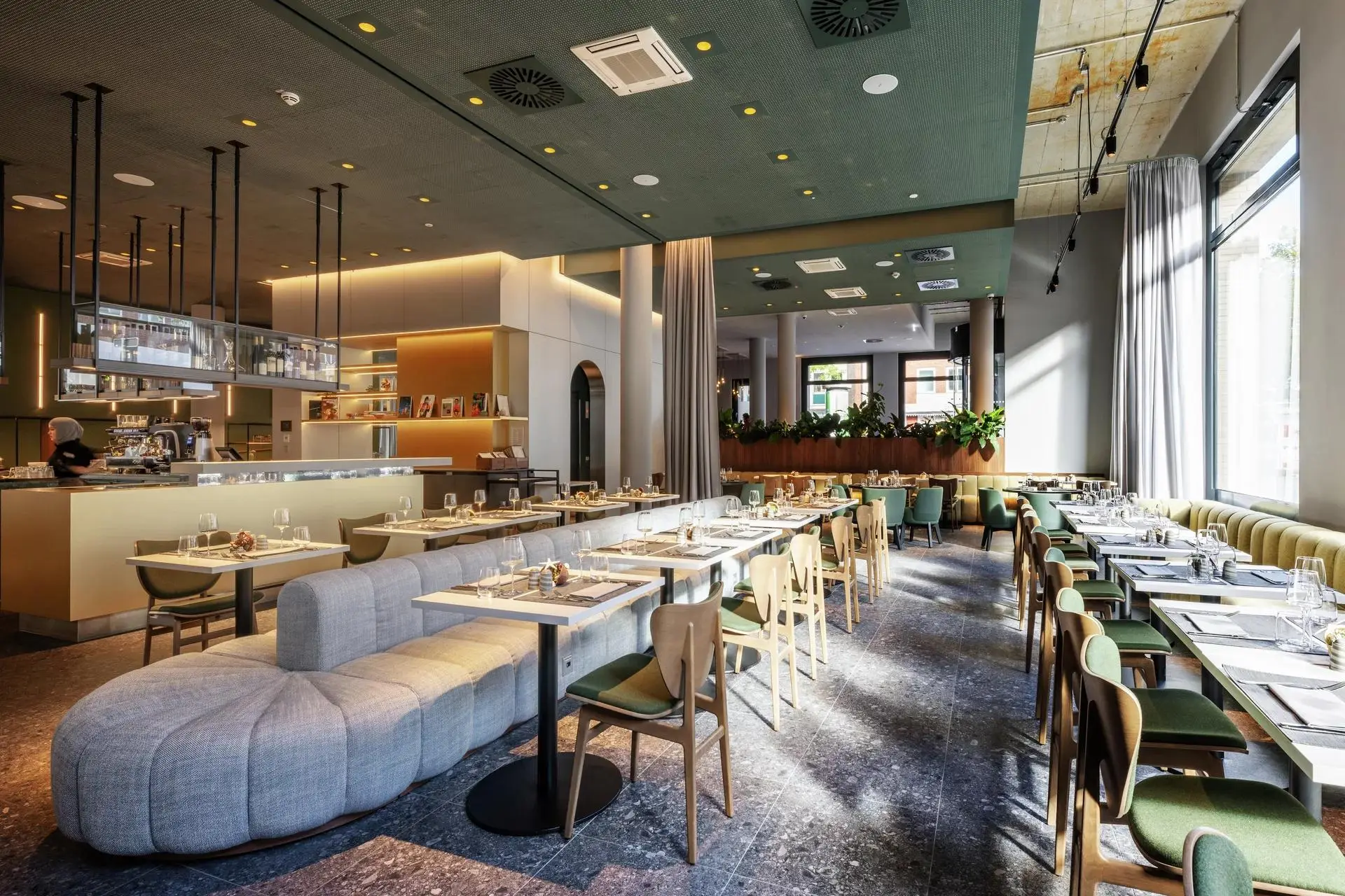 Bright modern restaurant dining room with set tables, green chairs, banquette seating, and a bar