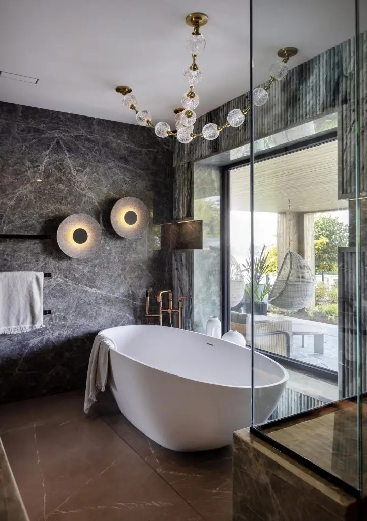Modern bathroom with a white freestanding bathtub, dark veined wall panels, decorative lighting, and a large window.