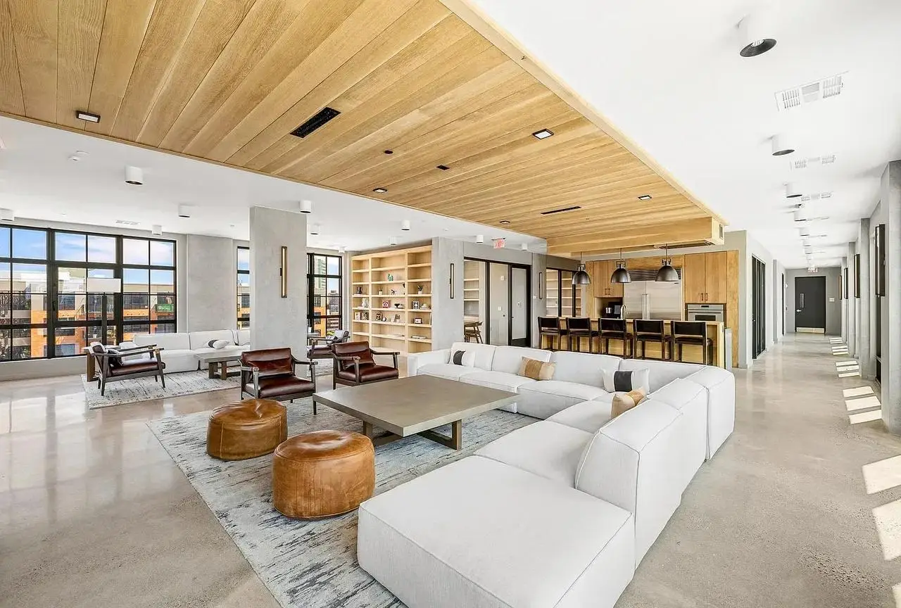 Bright modern lounge with white sectional sofas, leather ottomans, a wood-paneled ceiling, and an open kitchen area.