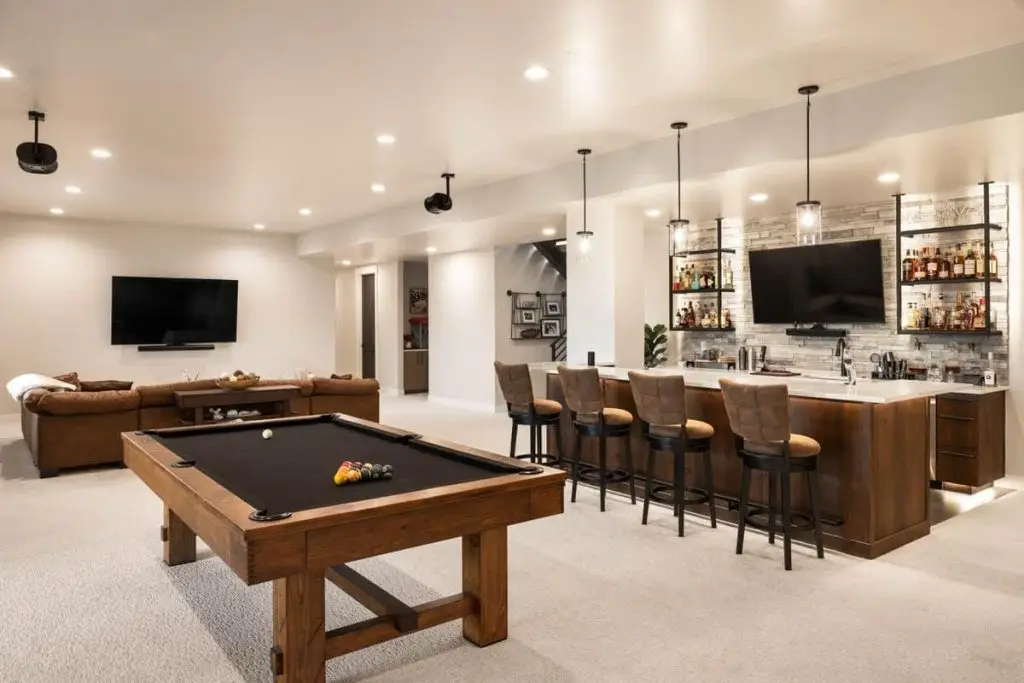 Finished basement with a pool table, sectional sofa, wall-mounted televisions, and a wood bar with stools and liquor shelves