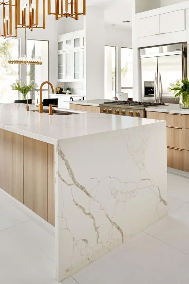 Bright modern kitchen with a large white veined island, wood cabinetry, brass fixtures, and stainless steel appliances