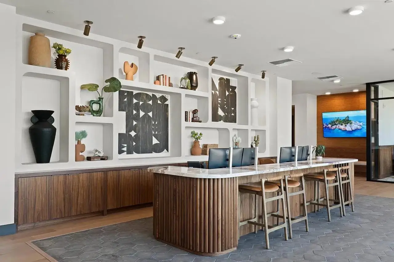 Modern reception lobby with a wood-paneled desk, computer monitors, stools, decorative shelving, and a wall-mounted television
